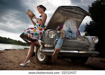 Pretty couple is trying to repair their old car using manual. Rural background.
