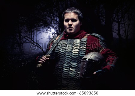 Medieval knight in the armor with the sword and helmet. Dark forest on the background.