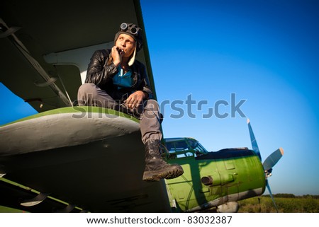 Smoking pilot in the helmet is sitting on the wing of a vintage plane.