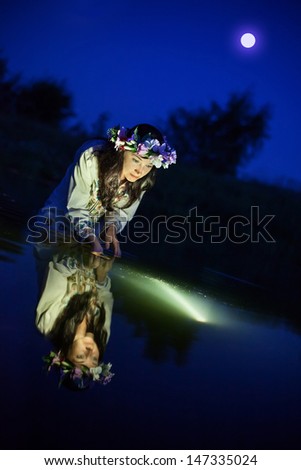 Beautiful woman in the night river dressed in the folk chemise and floral wreath