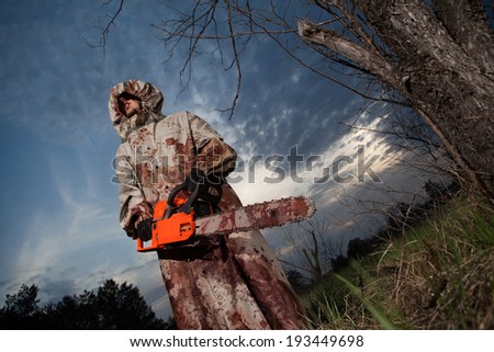 Maniac with the chainsaw dressed in a dirty bloody raincoat. Sunset forest on the background