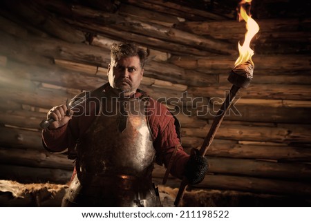 Medieval knight in the armor with the sword and flame. Wooden wall on the background.