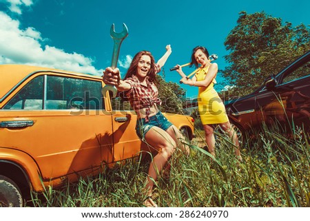 Cheerful woman mechanic is repairing a car with the wrench another woman is watching.