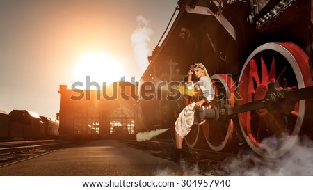 Woman in the vintage dress is sitting on the locomotive\'s wheel. Station on the background. Artistic lens flares are the part of composition.
