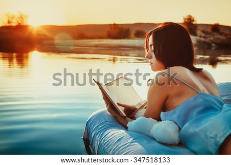 Pretty woman is laying on the floating bed and reading a book. Summer sunset on the background.