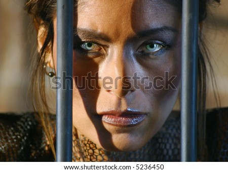 A woman with striking eyes stares out between the bars of a jail cell.