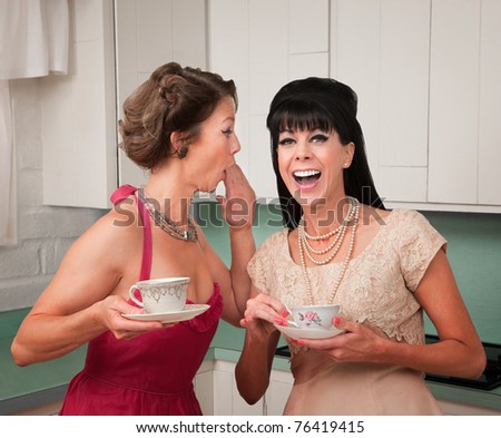 Caucasian woman whispers joke to friend in her kitchen