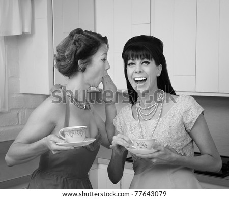 Caucasian woman whispers joke to friend in her kitchen