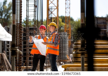 Construction manager and engineer dressed in orange work vests and hard helmets explore construction documentation on the building site near the steel frames