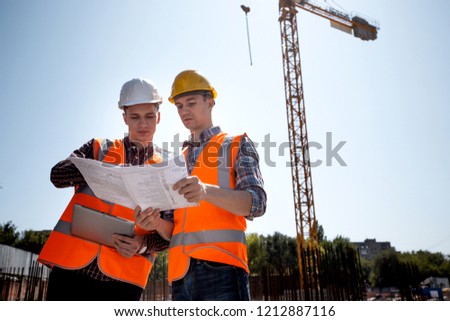 Architect  and construction manager dressed in orange work vests and helmets discuss documentation on the open air building site next to the crane