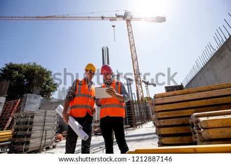 Civil architect  and construction manager  dressed  in orange work vests and  helmets discuss  a building project on the mobile tablet on the open building site next to the crane