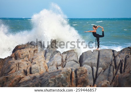 girl is doing yoga, standing in Lord of the Dance pose (Natarajasana) on rocks near crashing wave