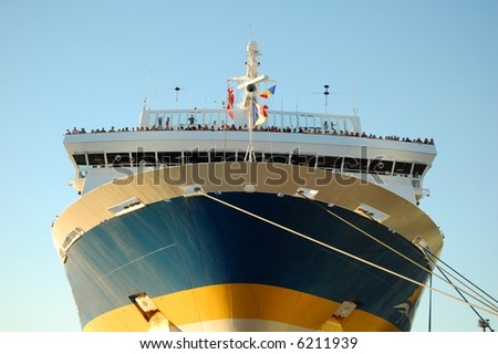 Picture of a passenger ship docked, about to leave the wharf. Passengers can be seen at the front, waiting to say goodbye to friends and families.