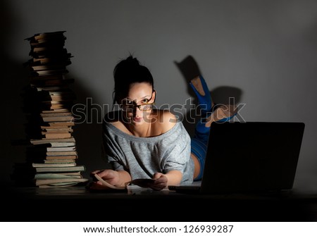 Beautiful girl student lying on the floor with laptop and books on a dark background.