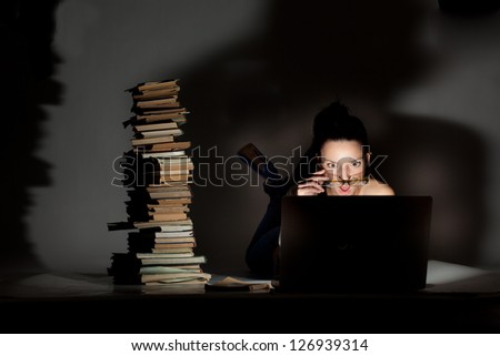 Beautiful girl student lying on the floor with laptop and books on a dark background.