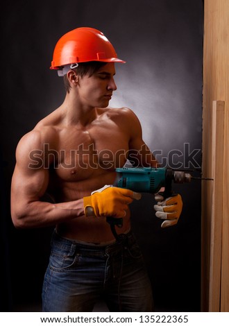 Muscular young worker in an orange helmet with an electric drill on a black background.