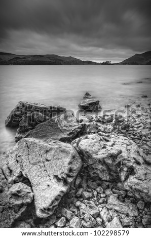 A black and white portrait format view over Derwent Water in the Lake District. A line of angular rocks in the foreground of the image receeds into the water while storm clouds rush by over head.