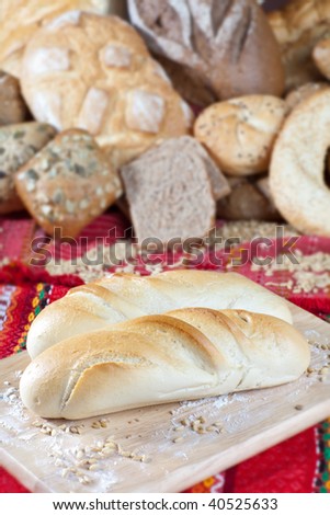 Two french rolls on wooden breadboard in front of heap of different types of bread