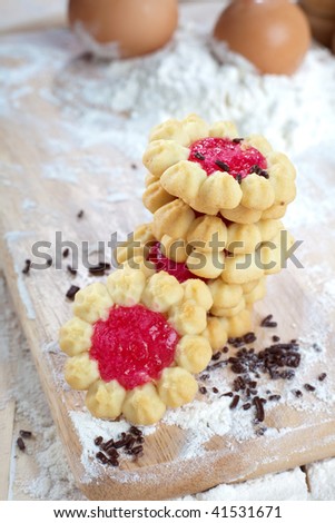 Stack of freshly made cookies on wooden bread board with two raw eggs and flour on the background