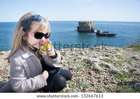Little Girl smelling flowers near Siracusa Faraglioni, in Sicily. The girl is wearing sunglasses and a coat: it is spring.