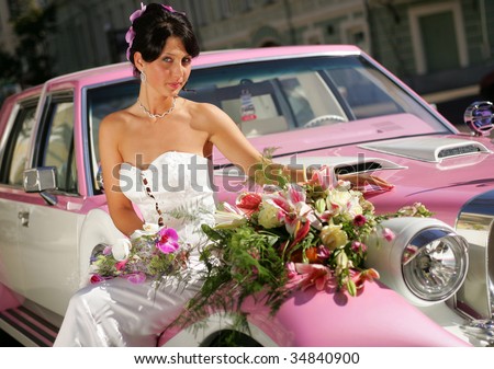 Pretty young adult bride sat on hood of pink wedding car limousine.