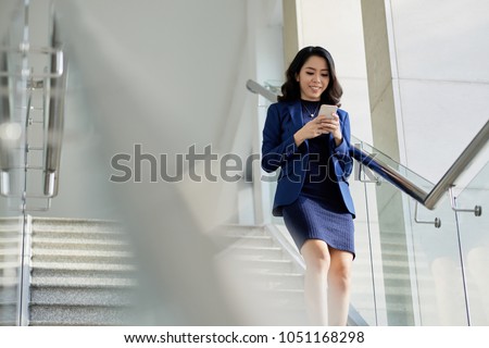 Smiling Asian manager in formalwear texting with friend on smartphone while going downstairs, portrait shot