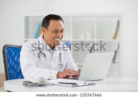 Portrait of mature doctor sitting in his office and typing on laptop