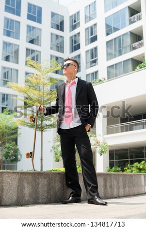 Trendy guy in business suit standing in urban environment holding a skateboard and listening to music