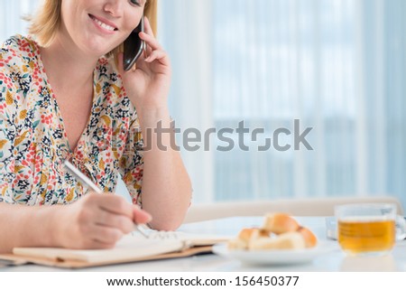 Image of a young woman talking by phone and making notes on the week-end on the foreground