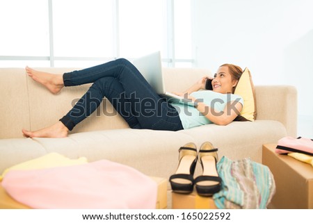 Image of a young online shopper with a computer and a phone lying on the sofa at home on the foreground