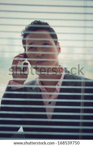 Vertical image of a young businessman talking by phone through the blinds on the foreground
