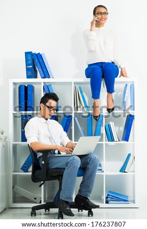 Vertical image of office workers where a man networking in the chair and a woman talking by phone seated on the cupboard