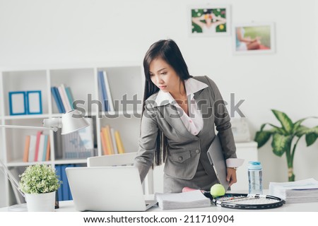 Serious Asian manager standing near desk and using laptop