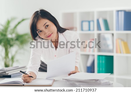 Pretty Asian manager talking on the phone and writing down in the office