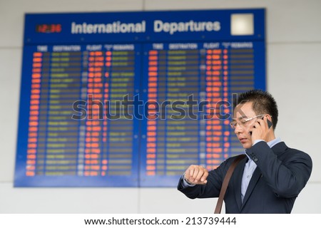Vietnamese businessman talking on the phone and checking the time while standing near flight timetable