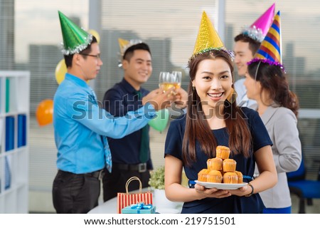 Portrait of beautiful manager holding a plate of cupcakes and her colleagues toasting in background