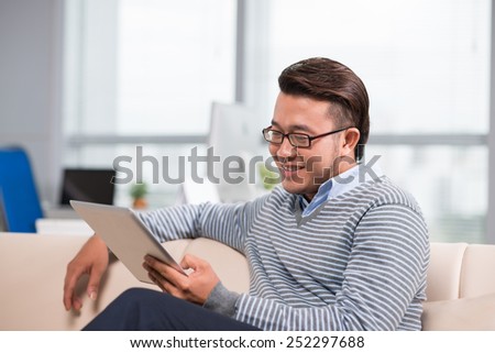 Asian man in glasses sitting on the sofa and reading something on the digital tablet