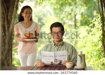 Aged Asian man reading a newspaper while his wife serving the table