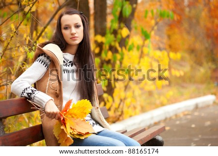 brunette woman sitting on bench and holding leaves in autumn park