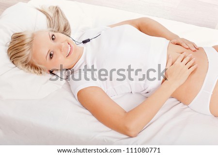 beautiful blonde woman laying on white bed and listening music and holding her hands on belly