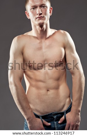 Handsome macho man posing shirtless to display his physique and muscles , upper body portrait on a dark studio background, looking at camera