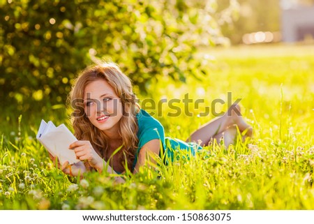 young blond woman reading a book lying on the grass in the shade, she is thinking about something