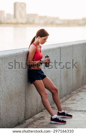 Female runner with bottled water tired from running standing near granite parapet