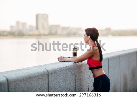 Female runner with bottled water tired from running standing near granite parapet, looking far away