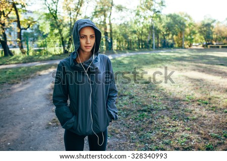 sporty woman wearing hood jacket and listening music outdoors in the park, cool toned image