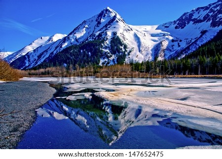 A Partially Frozen Lake With Mountain Range Reflected In The Partially Frozen Waters Of A Lake In The Great Alaskan Wilderness. A Beautiful Landscape Of Blue Sky, Trees, Rock, Snow, Water And Ice.