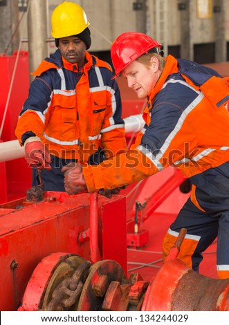 European deck officer wearing orange overall and red hardhat working together with African sailor wearing orange overall and yellow hardhat on red deck of a ship during mooring operation