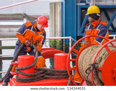 European deck officer wearing orange overall and red hardhat working with ropes together with African sailor wearing orange overall and yellow hardhat on red deck of a ship during mooring operation