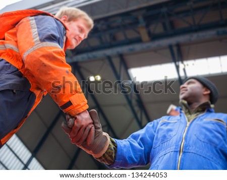Smiling European deck officer wearing orange overall and working gloves friendly shakes a hand of smiling African motorman wearing blue overall on red deck of a ship