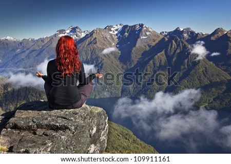 Red haired woman meditating in yoga position overlooking mountains and lake with white clouds.